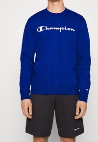 Modrý bavlněný svetr se dlouhými rukávy, kulatým výstřihem a bílým logem "Champion" přes hruď. V kombinaci s černými kraťasy.