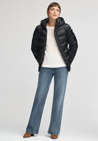 Czarna pikowana kurtka puchowa z kapturem, z przeszyciami i bocznymi kieszeniami, zestawiona z jasnym swetrem, szerokimi dżinsami i beżowymi botkami za kostkę.