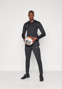 Schwarzer Sportanzug mit weißen Akzenten, langen Ärmeln und Reißverschluss, getragen von einer Person, die einen Fußball hält. Schwarze Schuhe.