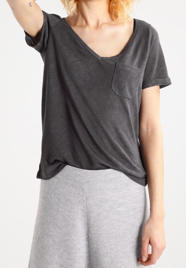 Personne portant un t-shirt ample gris foncé à col en V avec une poche poitrine et un pantalon texturé gris clair sur un fond uni.