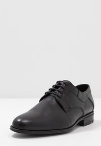 Chaussure habillée en cuir noir avec une texture perforée, bout rond, lacets et un petit talon. Présente une finition lisse et des accents de design minimalistes.