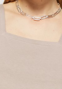 Béžová žebrovaná tílko se čtvercovým výstřihem, doplněné stříbrným řetízkem s obdélníkovými články a strukturovaným povrchem.