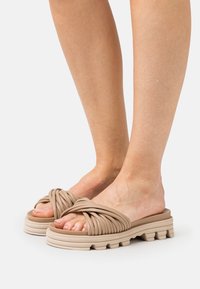 Sandales à glissière beige nouées avec une semelle épaisse et texturée, présentant des détails en relief, mettant en valeur un matériau doux et flexible pour le confort.