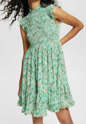 Vrouw draagt een mouwloze groene jurk met bloemenprint, gerimpelde zoom en geplooide lijfje, poseert met één hand op haar heup.