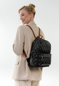 Hymyilevä nainen beigenvärisessä puvussa, jolla on musta kuvioitu reppu ruskeilla remmeillä, poseeraa sivuttain valkoista taustaa vasten.