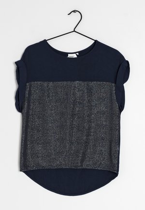 Blouse bleu marine à manches courtes avec manches retroussées et panneau argenté métallisé texturé à l'avant, suspendue sur un cintre noir.