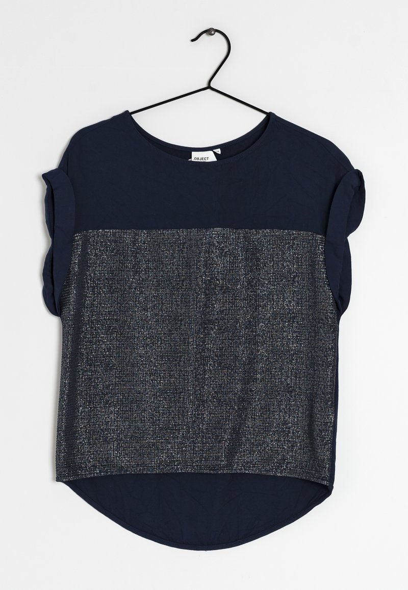 Blouse bleu marine à manches courtes avec manches retroussées et panneau argenté métallisé texturé à l'avant, suspendue sur un cintre noir.