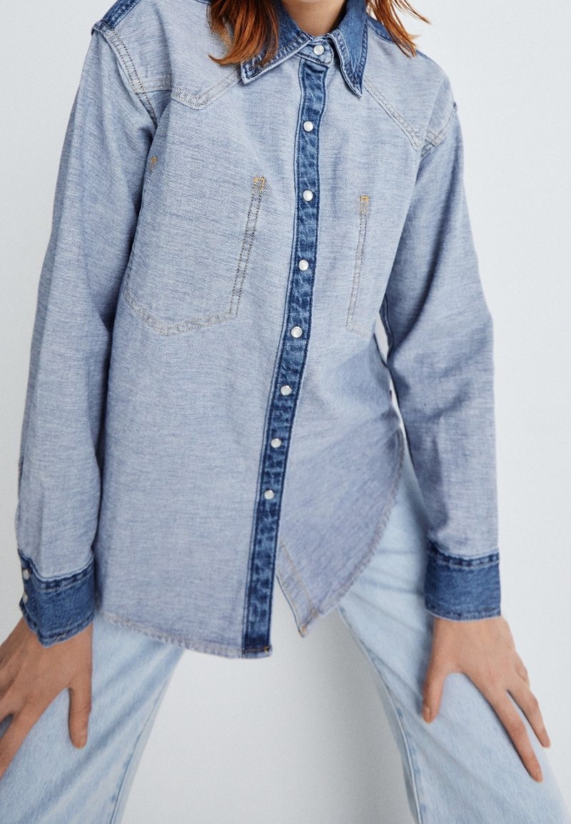 Femme portant une chemise en denim bleu clair avec des bordures bleu foncé et des boutons-pression, assortie à un jeans bleu clair, les mains reposant sur les cuisses.