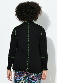 Zwart top met lange mouwen en gerimpelde groene bies langs de naden, met een hoge hals en aansluitende manchetten, gedragen met kleurrijke, met patronen bedrukte broek.