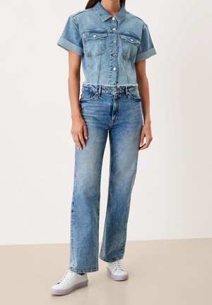 Vrouw draagt een kort mouw blauw denim overhemd met knopen, hoge taille rechte blauwe spijkerbroek en witte sneakers met lichtpaarse zolen.