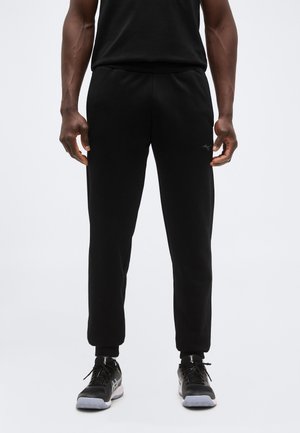 Άνδρας που φοράει μαύρο παντελόνι φόρμας και μαύρα αθλητικά παπούτσια, στέκεται μπροστά σε μια απλή φόντο.