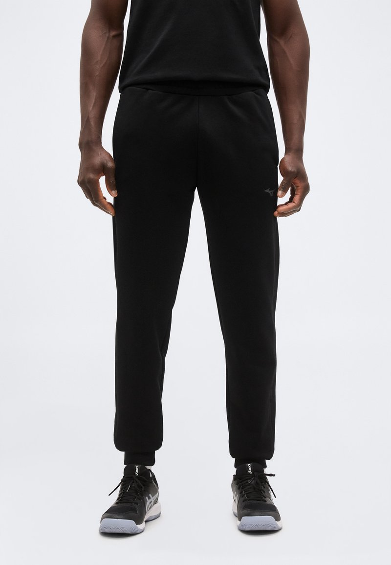 Άνδρας που φοράει μαύρο παντελόνι φόρμας και μαύρα αθλητικά παπούτσια, στέκεται μπροστά σε μια απλή φόντο.