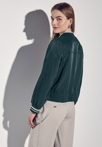 Veste bomber en vert foncé avec une texture douce, des poignets élastiques avec des rayures blanches, et une taille ajustée. Comprend une poche latérale.