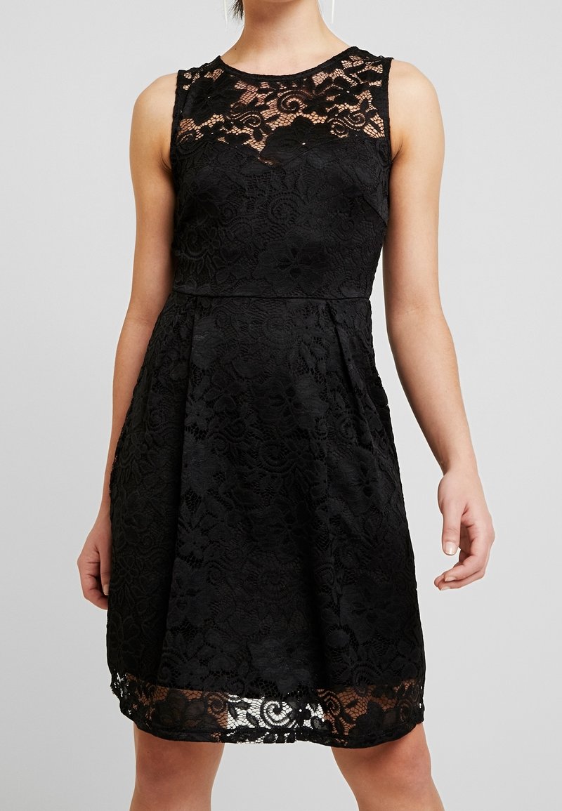 Robe noire sans manches en dentelle avec motifs floraux, taille ajustée, dentelle transparente à l'encolure et à l'ourlet, portée par une personne debout, les bras détendus.