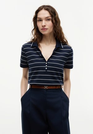 Γυναίκα με μακριά σγουρά μαλλιά που φοράει μπλε σκούρο πόλο μπλουζάκι με άσπρες οριζόντιες ρίγες και μπλε σκούρο παντελόνι ψηλής μέσης.