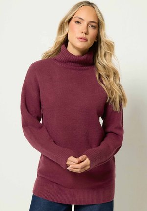 Femme aux longs cheveux blonds portant un pull à col roulé bordeaux et un jean bleu, debout les mains jointes devant.