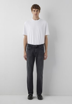 Junger Mann, der nach vorne steht, trägt ein schlichtes weißes T-Shirt, eng anliegende schwarze Jeans und schwarze Schuhe vor einem grauen Wandhintergrund.