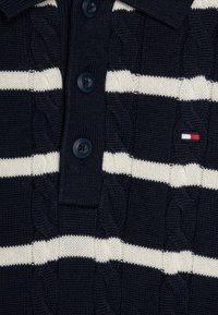 Marineblå strikket polo med kremfargede horisontale striper, med et teksturert kabelmønster, tre knappestolper og en liten logo i rødt, hvitt og blått.