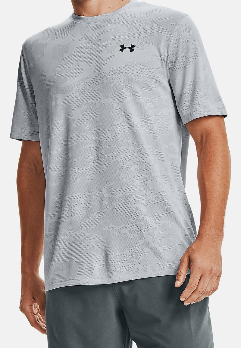 Harmaa lyhythihainen urheilupaita, jossa on teksturoitu maastokuvio ja musta logo vasemmassa rinnassa. Kevyt ja hengittävä kangas.