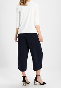 Weißes 3/4-Ärmel-Top kombiniert mit navy-farbenen, verkürzten Hosen. Schwarze High Heels mit Punkten und Knöchelriemen. Glatte Stofftextur.