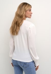 Blusa bianca con texture, maniche a palloncino lunghe, polsini elastici e scollatura tonda, abbinata a jeans azzurri aderenti.