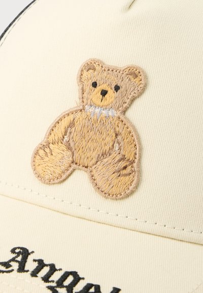 Κεντημένη ανοιχτόχρωμη αρκουδίτσα με μαύρα μάτια και μύτη πάνω σε υφασμάτινο κρεμ φόντο, πάνω από κεντημένο μαύρο κείμενο σε καπέλο ή ρούχο.