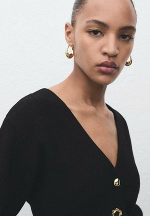 Black garment with a deep V-neckline, featuring a gold button detail. The model wears gold hoop earrings, showcasing a smooth texture and fitted design.
