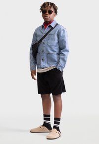 Denim jacket, red shirt, black shorts, beige slip-on shoes, and striped socks; accessories include black crossbody bag and sunglasses.