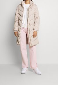 Abrigo acolchado beige con diseño de flechas, cuello alto y cierre de cremallera, combinado con pantalones de pierna ancha rosa y zapatillas blancas.