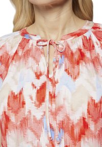 Lätt blus med ett abstrakt mönster i röd, blå och krämfärgad färg, som drar uppmärksamhet till halsområdet med en knytning och mjuk textur.