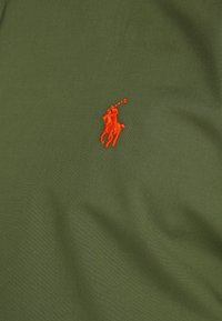 Olivově zelené polo tričko vyrobené z hladké bavlněné látky, s malým červeným vyšívaným logem koně a jezdce na hrudi.