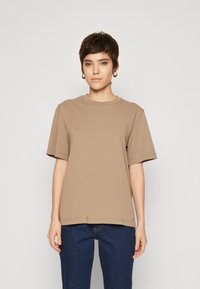 Braun Baumwoll-T-Shirt mit Rundhalsausschnitt und kurzen Ärmeln. Hat ein dezentes Logo auf der Vorderseite, kombiniert mit dunkelblauen Jeans.