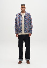 Bunte gemusterte Jacke mit blauen, orangefarbenen und grünen Blumenmotiven, getragen über einem cremefarbenen Hemd, kombiniert mit dunklen Jeans und braunen Schuhen.