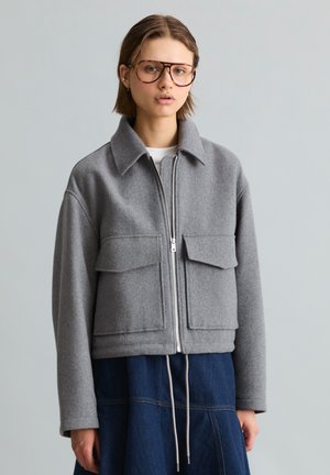 Jonge vrouw die een oversized grijze jas met rits en grote voorzakken en een blauwe rok draagt, staand tegen een effen lichte achtergrond.