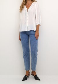 Witte, lichte blouse met driekwart pofmouwen, knoopsluiting aan de voorkant en decoratieve stiksels. Gecombineerd met lichtblauwe jeans en zwarte hakken.