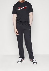 Svart Nike-t-shirt med röd och vit "AIR"-logga, kombinerad med svarta sweatpants med en liten vit Nike-swoosh och svarta och vita sneakers.