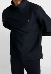 Granatowy sweter z dzianiny z wysokim kołnierzem, charakteryzujący się fakturowanym materiałem i małym złotym logo na lewej piersi. Bocznie rozcięcia na dole.