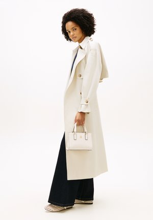 Frau mit lockigem Haar, die einen beigen Trenchcoat und dunkle Hosen mit weitem Bein trägt, hält eine kleine weiße, strukturierte Handtasche und steht im Profil.