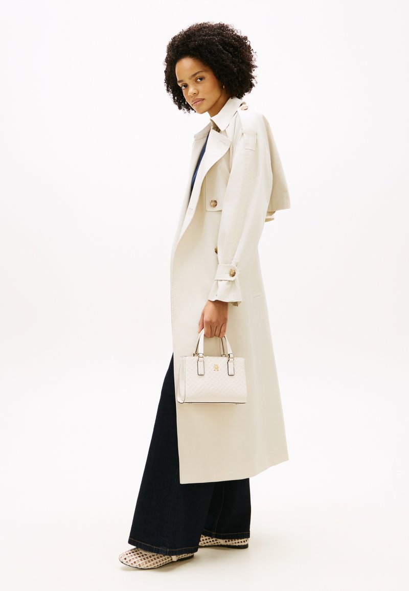 Γυναίκα με σγουρά μαλλιά, ντυμένη με μπεζ καμπαρντίνα και σκούρο φαρδύ παντελόνι, κρατώντας μια μικρή λευκή τσάντα με υφή, όρθια σε προφίλ.