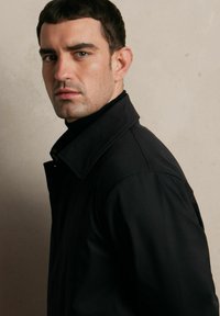 Czarna kurtka z kołnierzem, wykonana z gładkiego, matowego materiału, z metalowym suwakiem z przodu i prostymi liniami, noszona z czarnym swetrem z wysokim kołnierzem pod spodem.