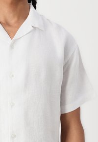 Chemise blanche en lin à manches courtes avec devant à boutons, texture douce et coupe décontractée, dotée d'un col classique.