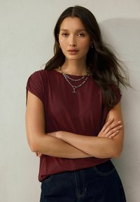 Burgundy t-shirt with short sleeves and a relaxed fit, paired with a dark denim bottom and layered silver chain necklaces.