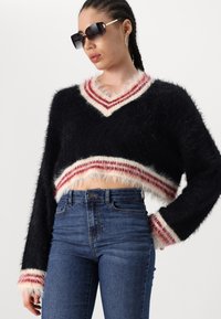Svart kortkort tröja med en fluffig textur; har en v-ringning med röda och vita randiga detaljer. Kombineras med högmidjade blå jeans.