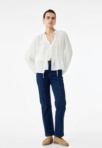 Camisa blanca de textura, con diseño peplum y botones. Combinada con jeans de pierna recta en azul oscuro y zapatos tipo slip-on en marrón claro.