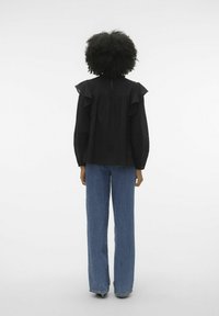 Blusa nera in tessuto strutturato con spalle arricciate e chiusura a bottoni sul retro, abbinata a jeans blu a gamba dritta.