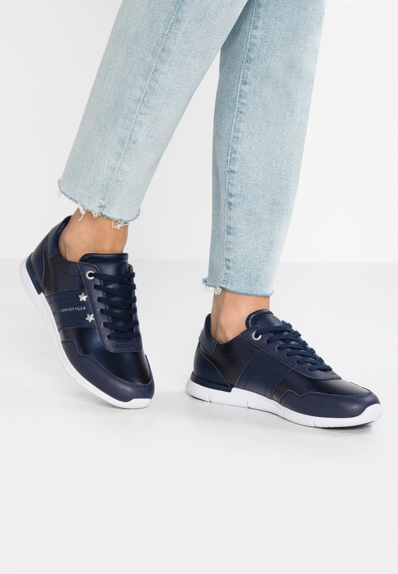Personne portant des baskets en cuir bleu marine avec semelles blanches et détails en forme d'étoile, associées à un jean bleu clair à ourlet effiloché.