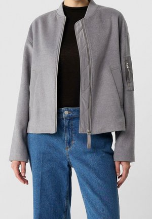 Persoon draagt een grijze bomberjack met rits over een zwart shirt, gecombineerd met een hoge taille blauwe spijkerbroek tegen een effen achtergrond.