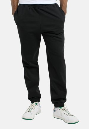 Mand iført sorte joggebukser med elastiske manchetter og hvide sneakers med grønne detaljer, stående med hænderne i lommerne.