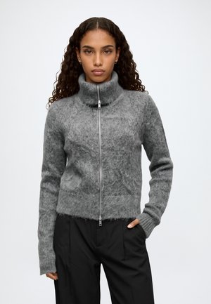 Femme aux longs cheveux bouclés portant un pull gris en peluche zippé et un pantalon noir, debout avec une main dans la poche sur un fond blanc.
