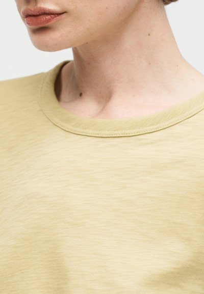 Κοντινό πλάνο ενός ατόμου που φοράει ένα ανοιχτό κίτρινο μπλουζάκι με στρογγυλή λαιμόκοψη, εμφανίζοντας το κάτω μέρος του προσώπου, τον λαιμό και το πάνω μέρος του στήθους σε απλό φόντο.